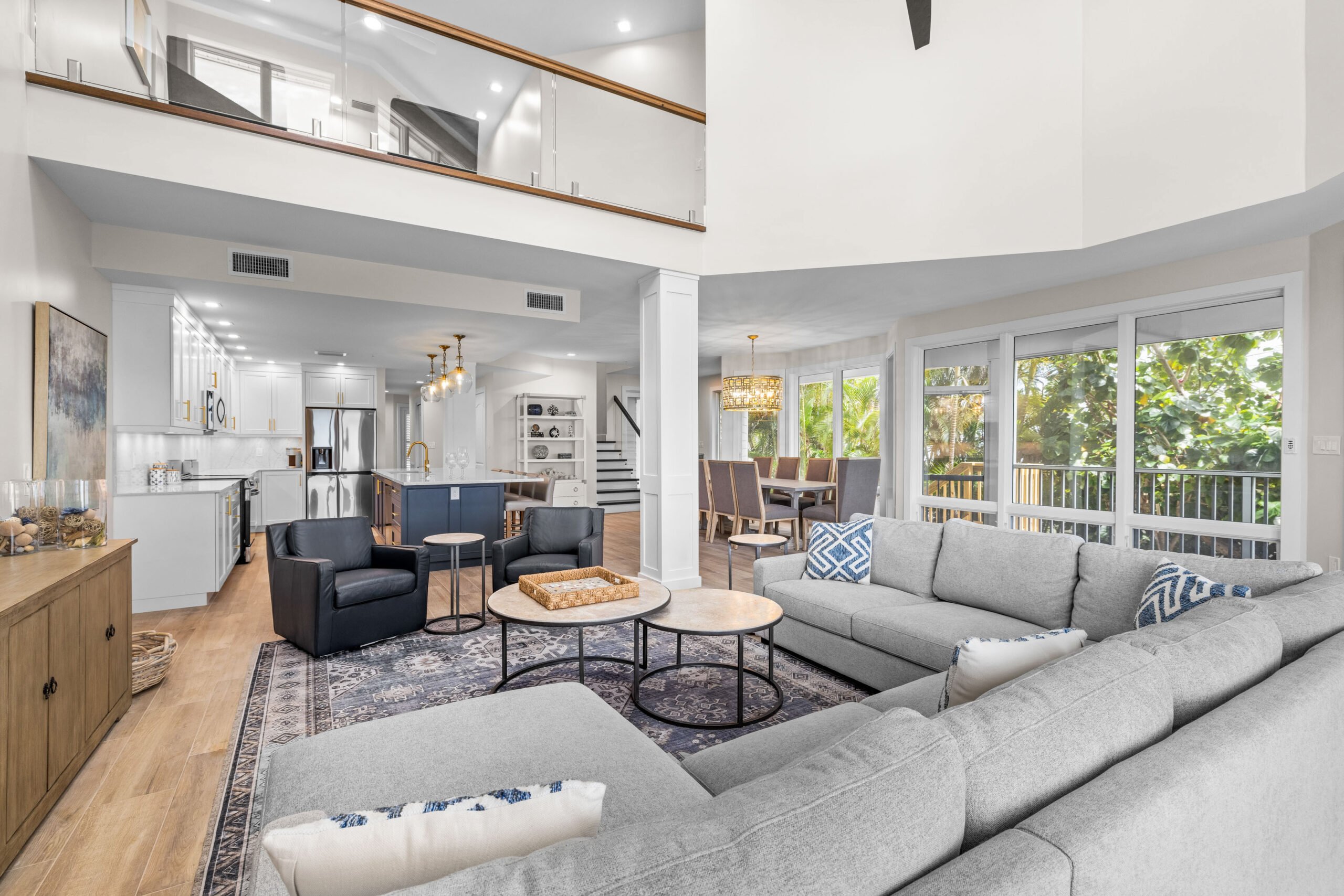 Gray sectional, two round tables, large windows, open kitchen and dining.