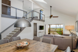 Wood table, gray chairs, sectional sofa, hanging light, stairs, ocean view.